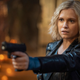 Clarke, a blond woman in a leather coat, points a gun at someone off screen.